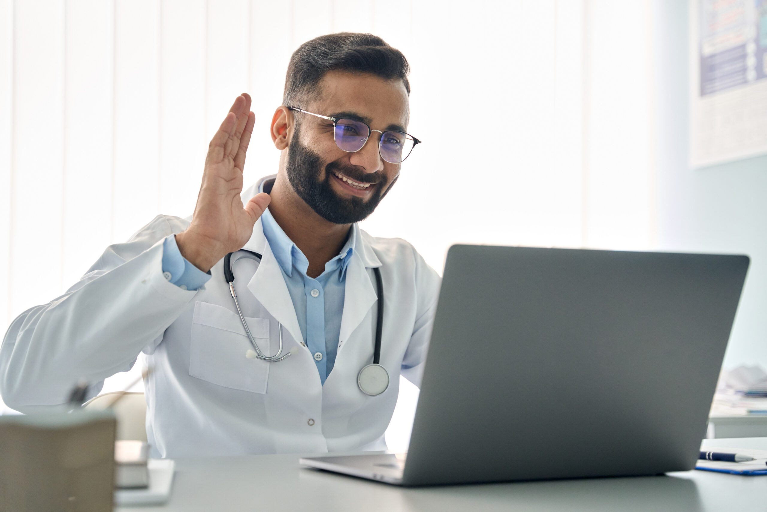Indian male doctor waving hand having virtual online consultation on laptop. - Recuro Health | Making Advanced Healthcare Accessible™